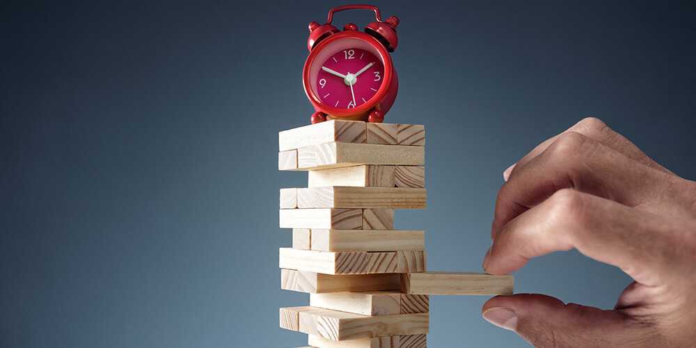 Photo of a hand removing a single block from a stack of Jenga tiles, while precariously balancing an alarm clock. This concept photo represents the balance of time and tricky discovery decisions.
