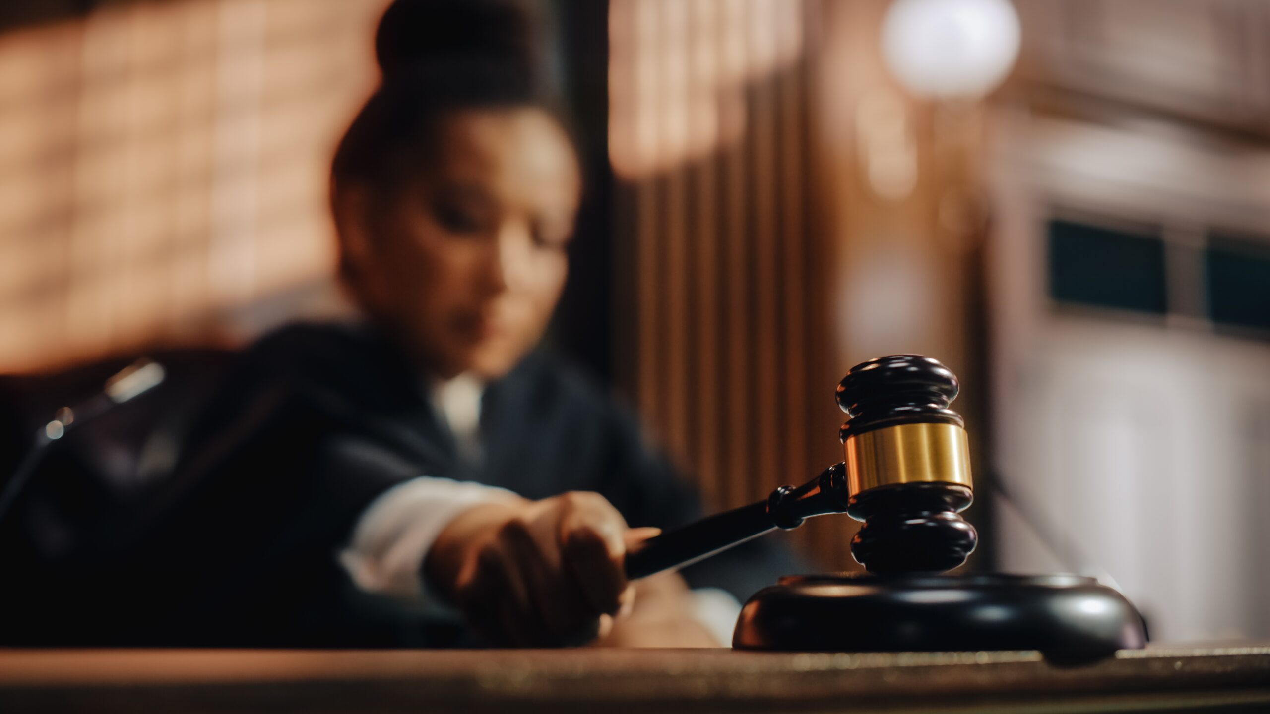 Focused image of a judge, partially visible and out of focus, about to strike a wooden gavel onto its base in a courtroom setting, with a warm glowing light in the background creating a serious atmosphere.