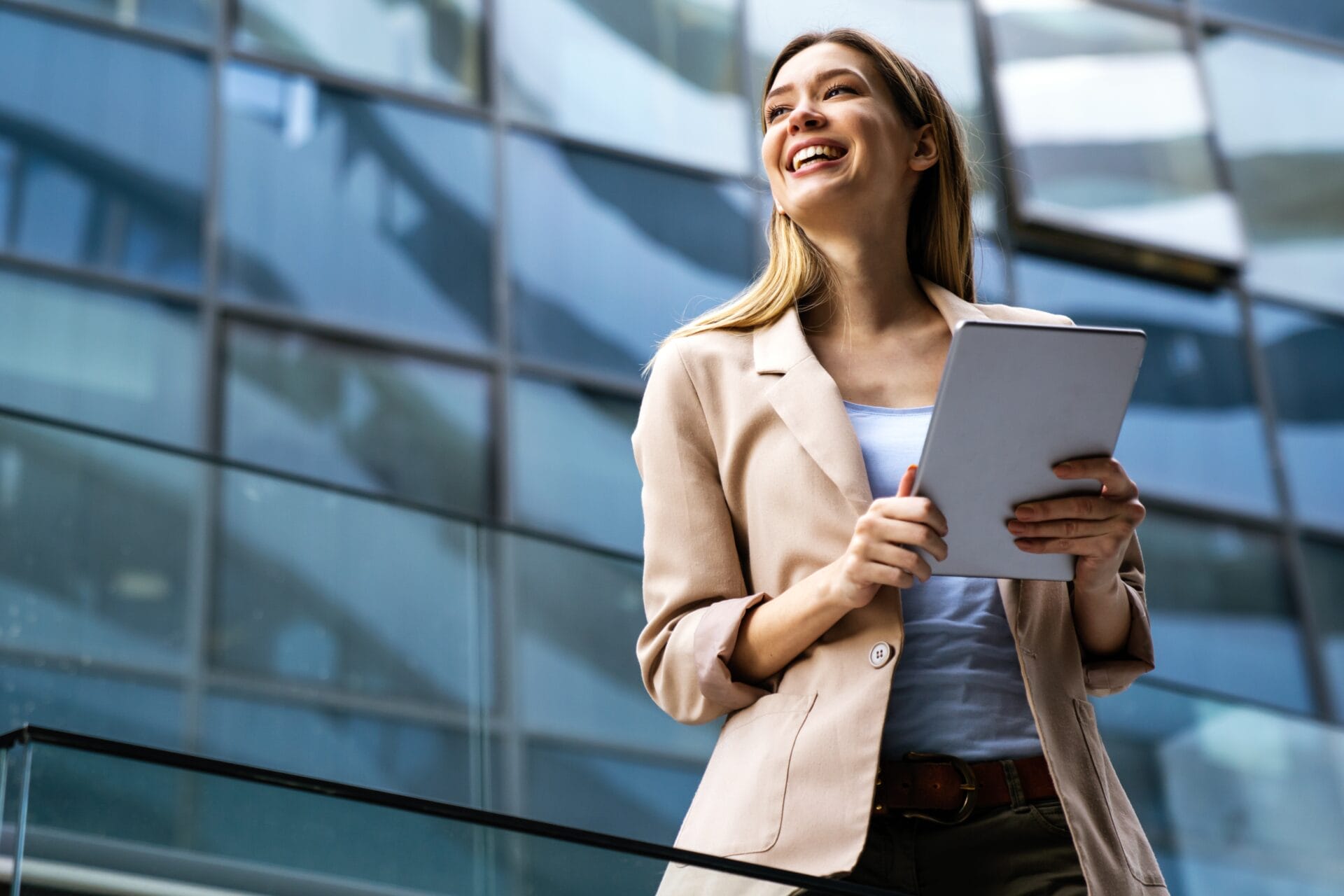 A professional woman with a joyful expression holding a tablet while standing outdoors. She's wearing a light beige blazer, a white top, and has her hair styled in loose waves. The background features a modern glass building, suggesting a corporate environment.