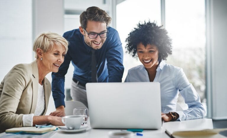 Three professionals collaborate around a laptop in a bright, modern office. An older woman with short hair smiles as she looks at the screen, alongside a younger man and woman who appear engaged and happy. The setting suggests a productive and supportive work environment, reflecting teamwork and technological interaction.