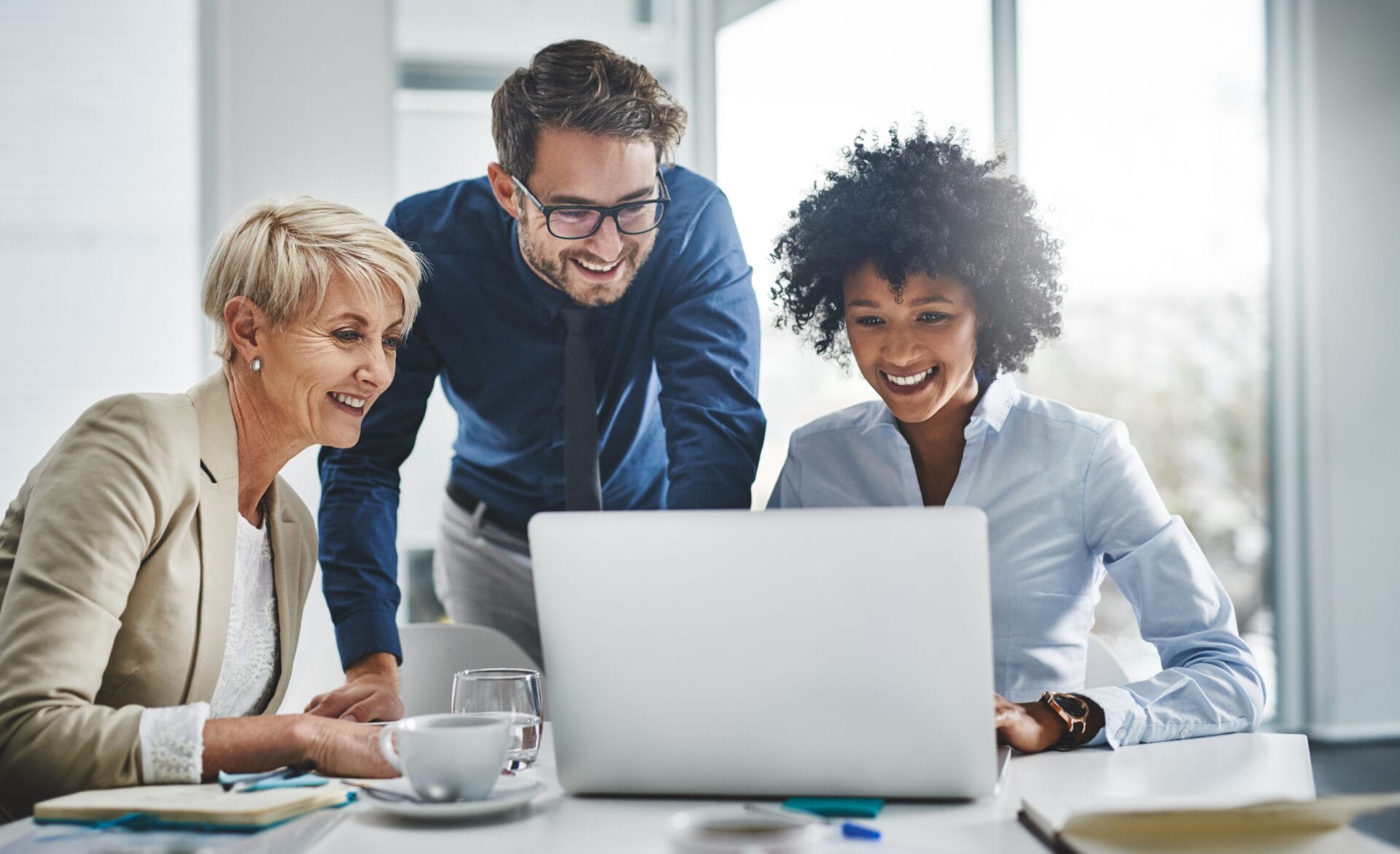 Three professionals collaborate around a laptop in a bright, modern office. An older woman with short hair smiles as she looks at the screen, alongside a younger man and woman who appear engaged and happy. The setting suggests a productive and supportive work environment, reflecting teamwork and technological interaction.